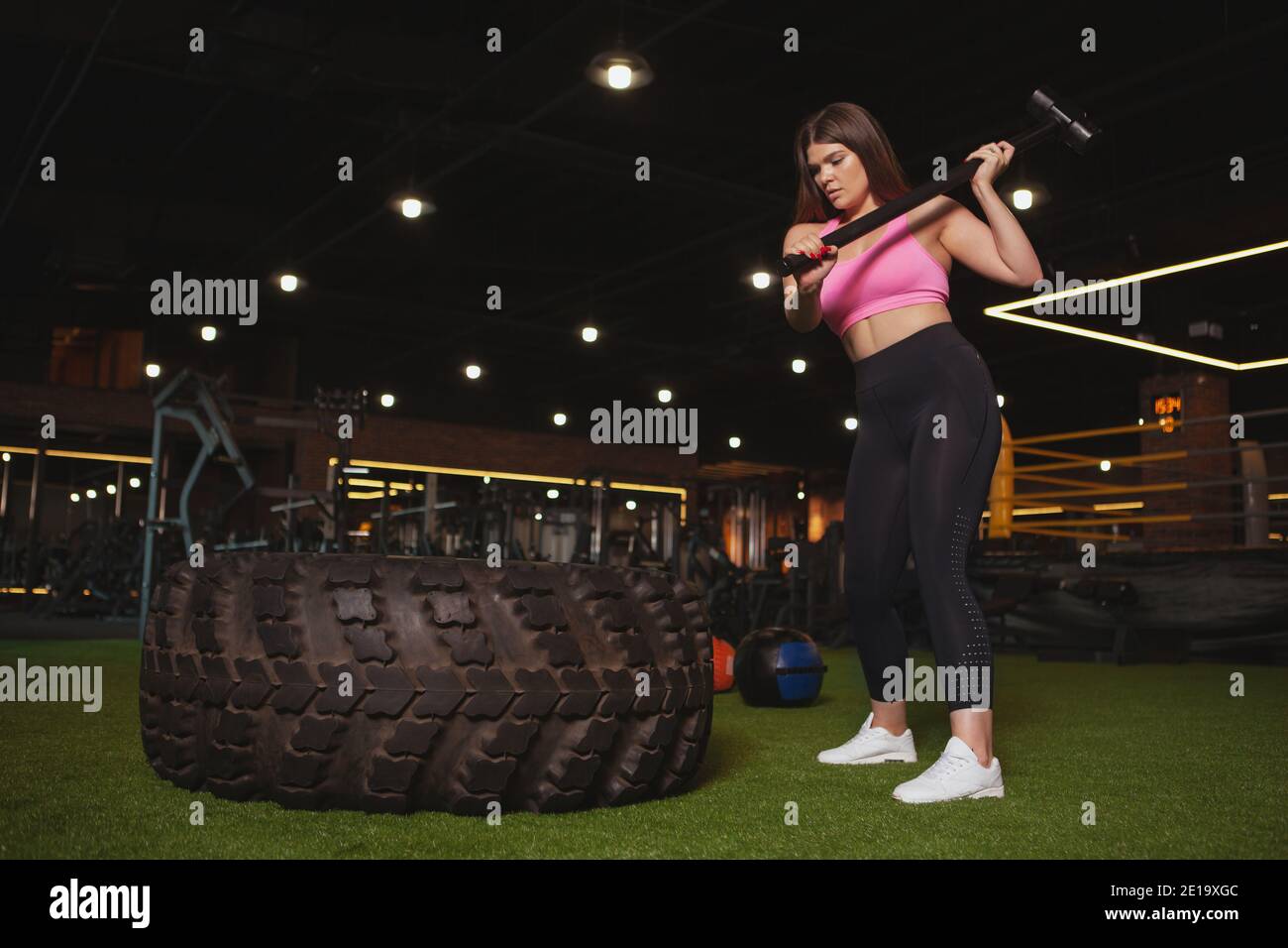 Prise de vue en longueur d'une jeune sportwoman de plus grande taille frappant un énorme pneu de roue avec un marteau en traîneau, espace de copie. Belle femme curvy faisant le travail crossfit Banque D'Images
