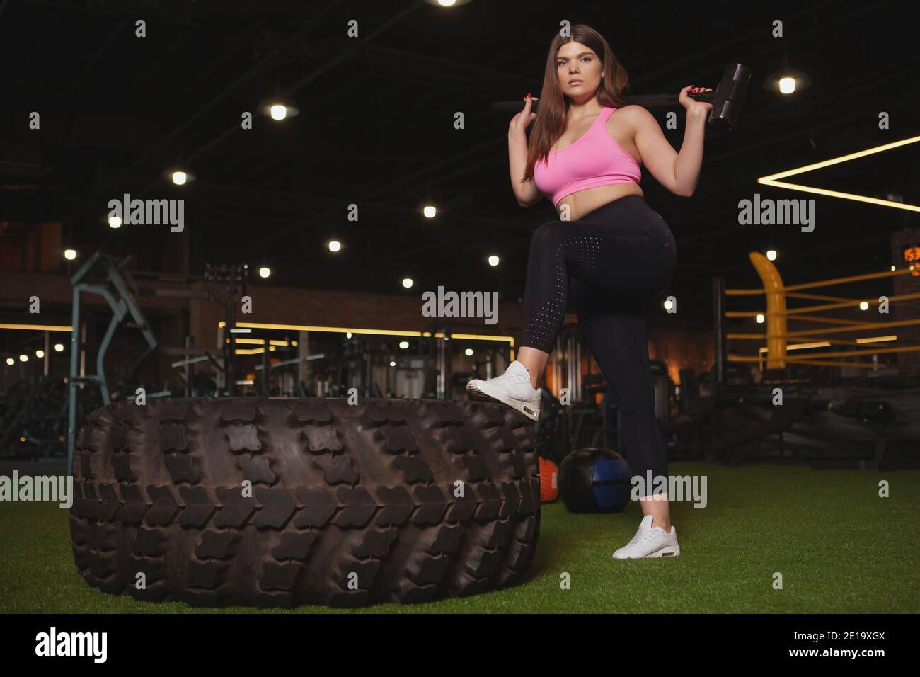 Prise de vue en basse angle d'une sportive de grande taille et belle et pleine de confiance, regardant avec force l'appareil photo, posant avec un marteau près de la roue lourde à la salle de gym, Banque D'Images