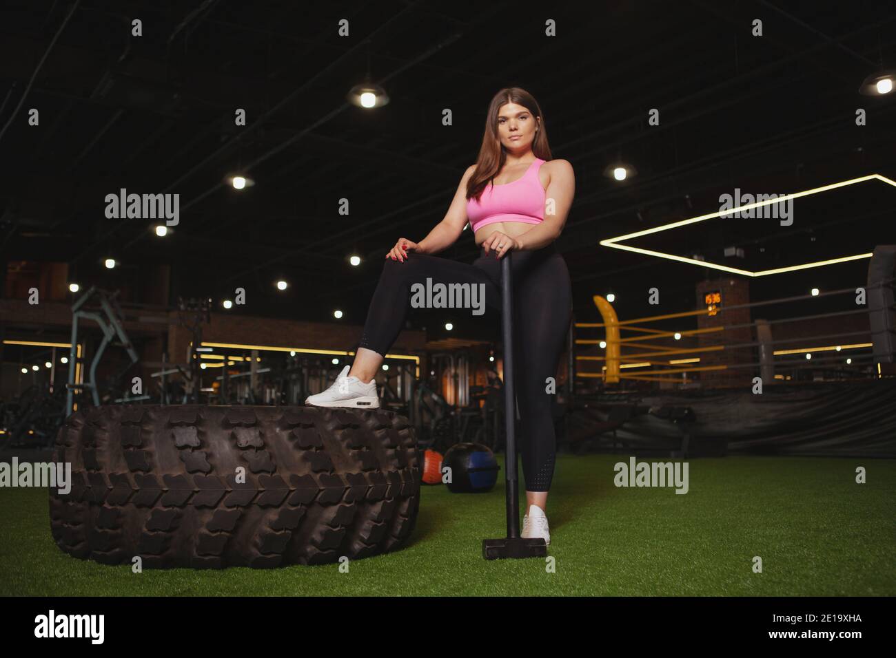 Jeune femme attrayante et de taille plus regardant avec confiance la caméra, se reposant après avoir fait de l'entraînement avec un marteau en traîneau et un énorme pneu de roue lourd. Gym, corps Banque D'Images