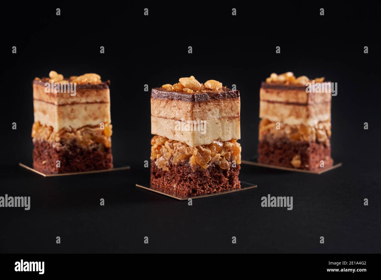 Vue avant des morceaux de dessert frais superposés avec biscuit et mousse isolés sur fond noir. Vue en gros plan de trois tranches carrées de gâteau au chocolat brun sucré avec dessus glacé et noix. Banque D'Images