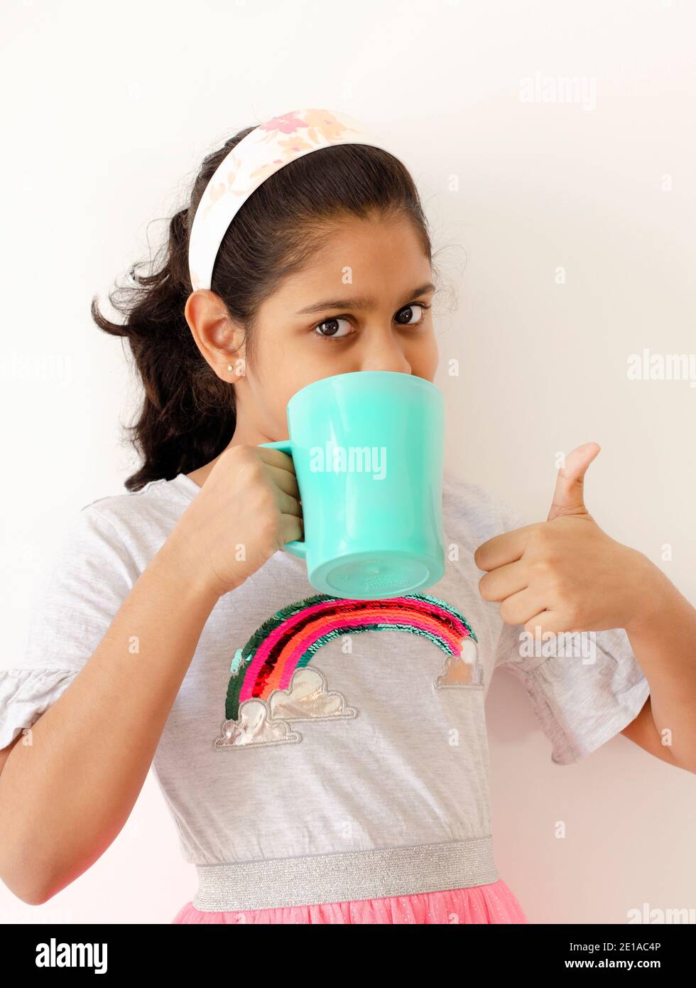 Portrait d'une adorable fille indienne buvant une tasse de vert bleuâtre et montrant les pouces vers le haut. Banque D'Images