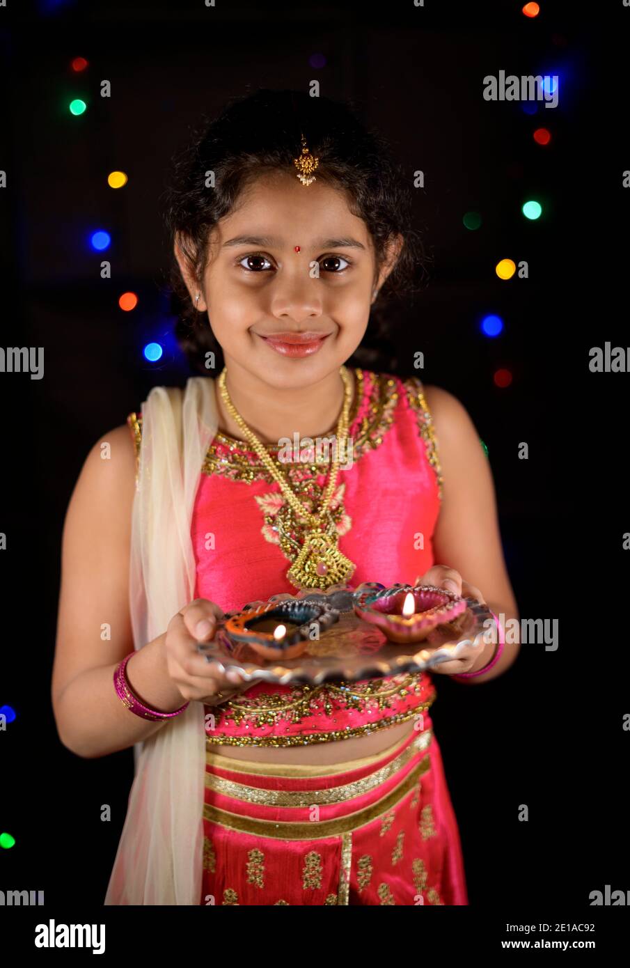 Une belle fille indienne souriante vêtue de vêtements ethniques avec des bijoux et tenant des lampes à huile pendant diwali la nuit avec des lumières sombres dans l'arrière-plan. Banque D'Images