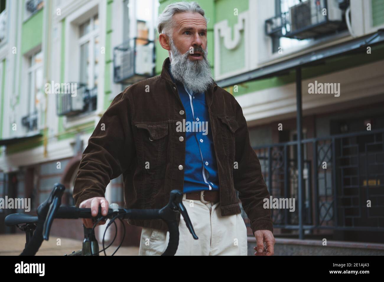 Beau barbu homme élégant avec cheveux gris marchant dans les rues de la ville avec son vélo Banque D'Images