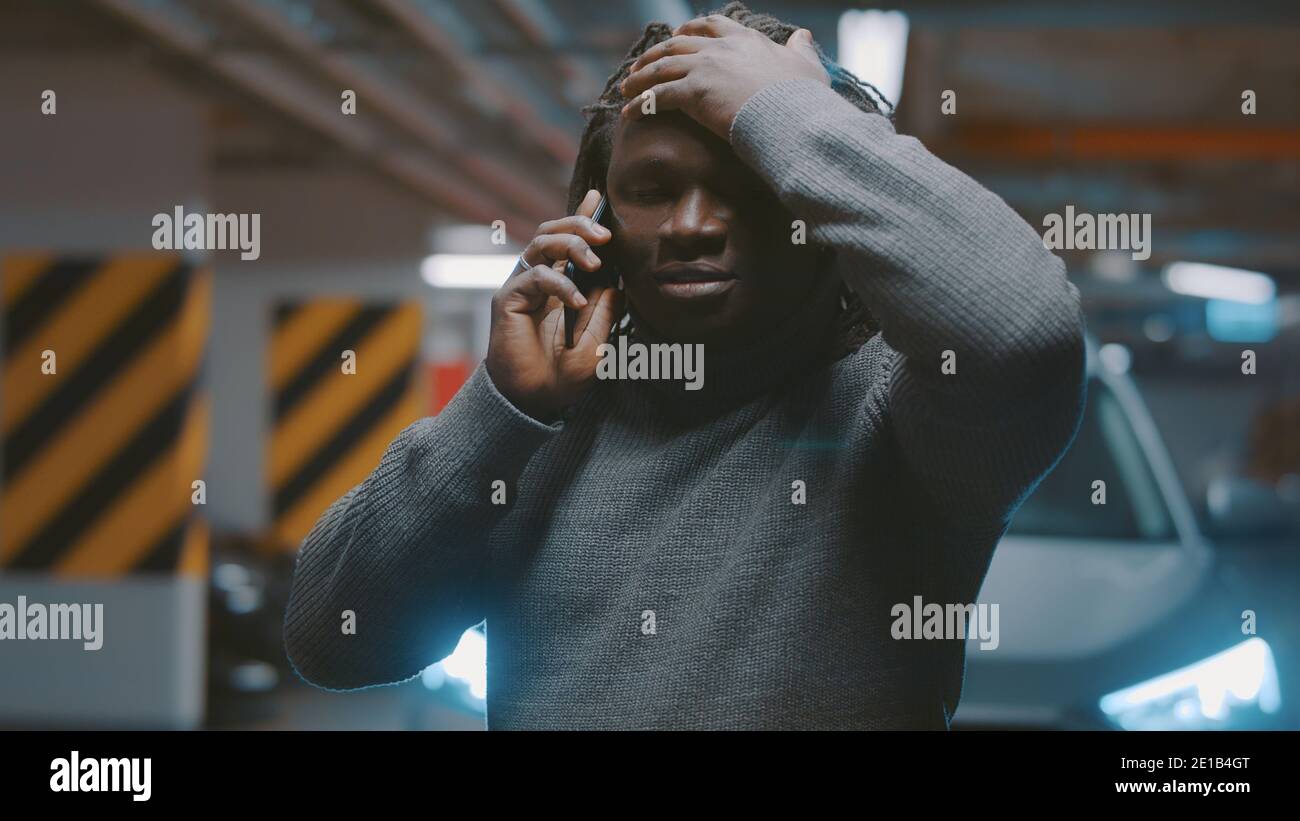 Contrarié afro-américain homme noir parlant sur le smartphone devant la voiture dans le garage. Photo de haute qualité Banque D'Images