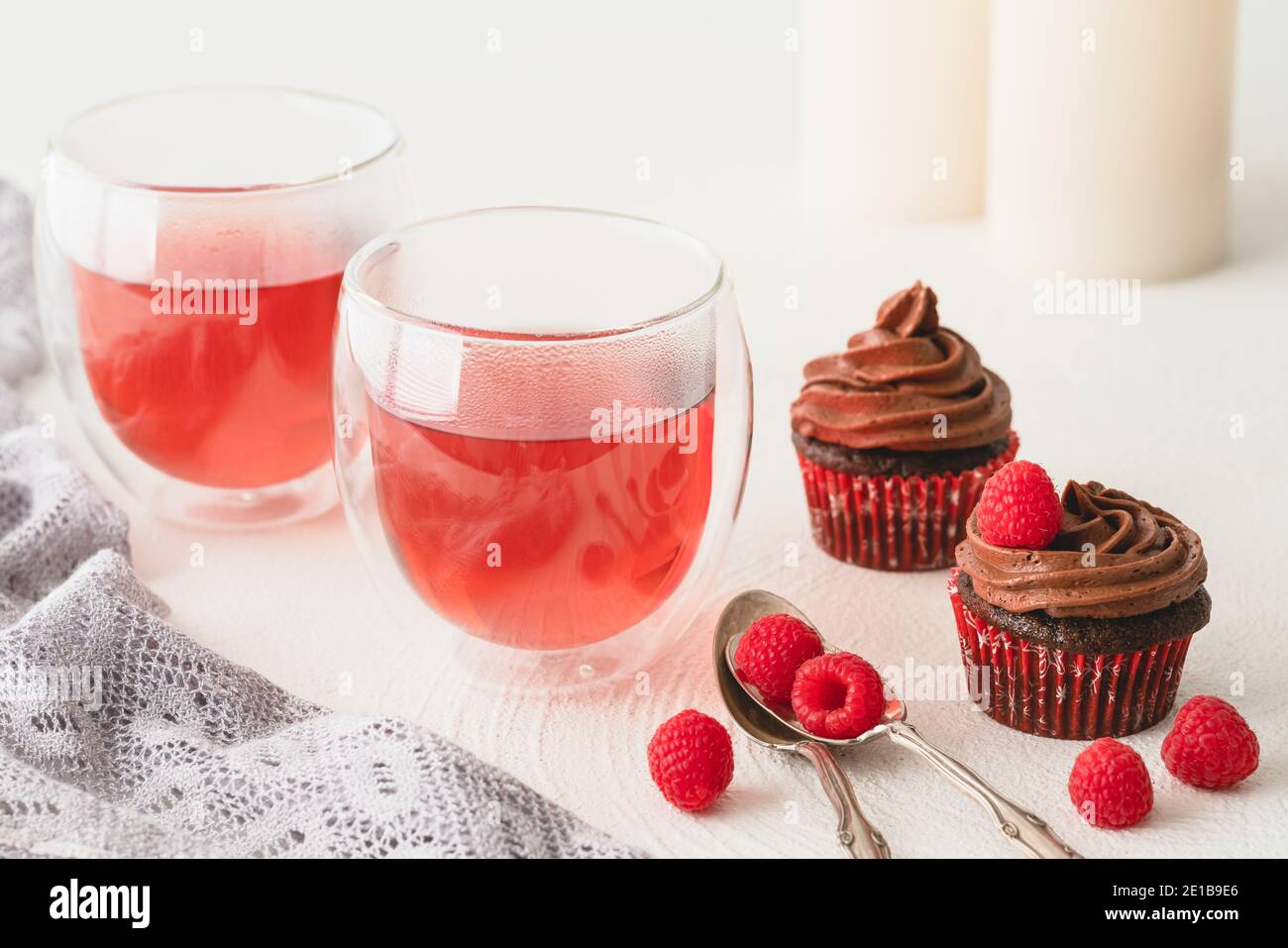 Thé à la framboise dans des tasses en verre et petits gâteaux au chocolat décorés de glaçage au chocolat et de framboises fraîches sur fond blanc. Délice sucré Banque D'Images