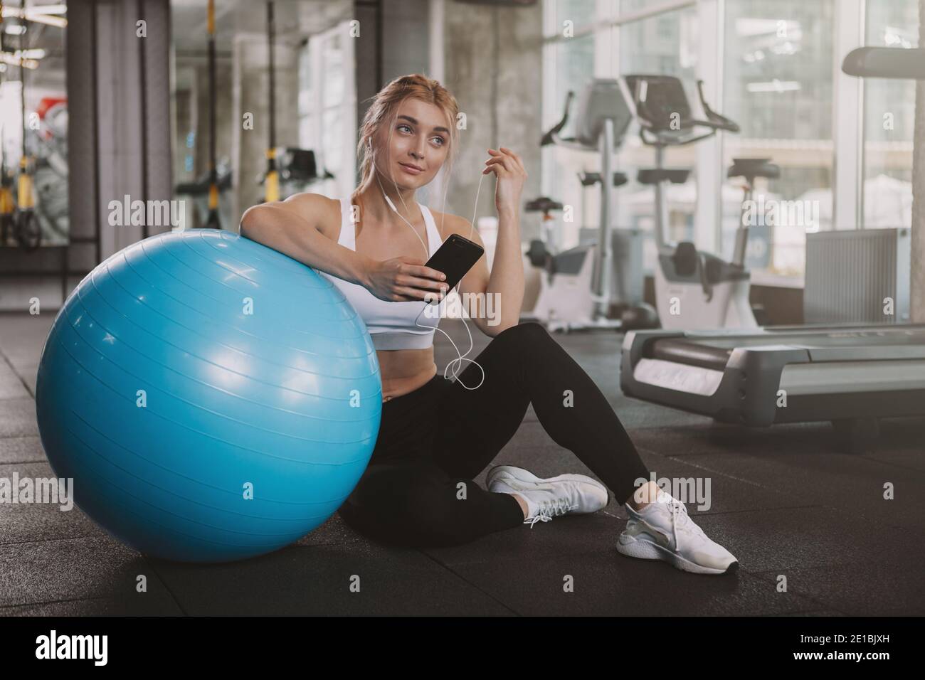 Magnifique heureux bonne santé jeune sportswoman utilisant son smartphone à la salle de gym, espace de copie. Belle femme sportive blonde aux cheveux se détendant après avoir fait de l'entraînement Banque D'Images
