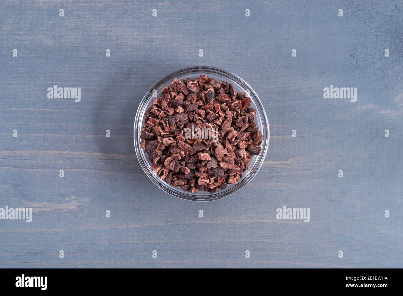 Vue de dessus d'un bol en verre rempli de nibs de cacao sur un plan d'examen en bois bleu pâle éclairé par la lumière naturelle. Banque D'Images