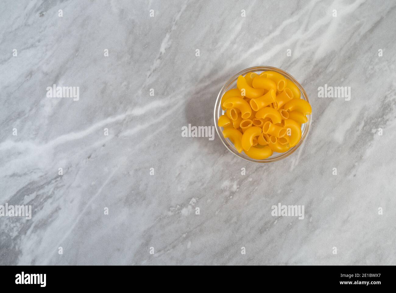 Macaroni au coude bio dans un bol en verre décalé sur un plan de travail gris éclairé par la lumière naturelle. Banque D'Images