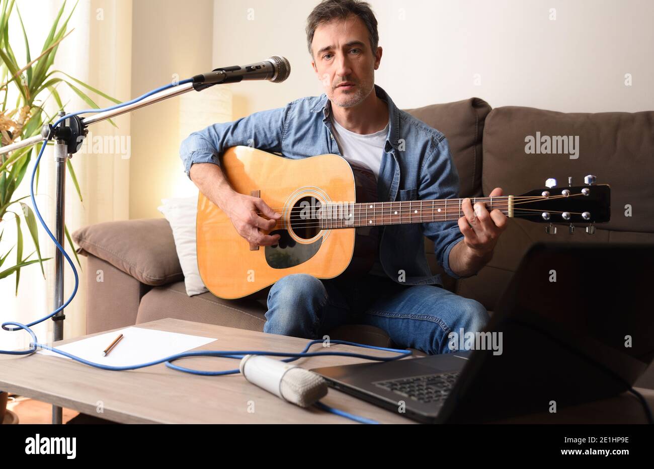 Homme jouant de la guitare acoustique à la maison sur un canapé avec équipement pour la musique avec microphone et ordinateur portable sur une table en bois. Banque D'Images