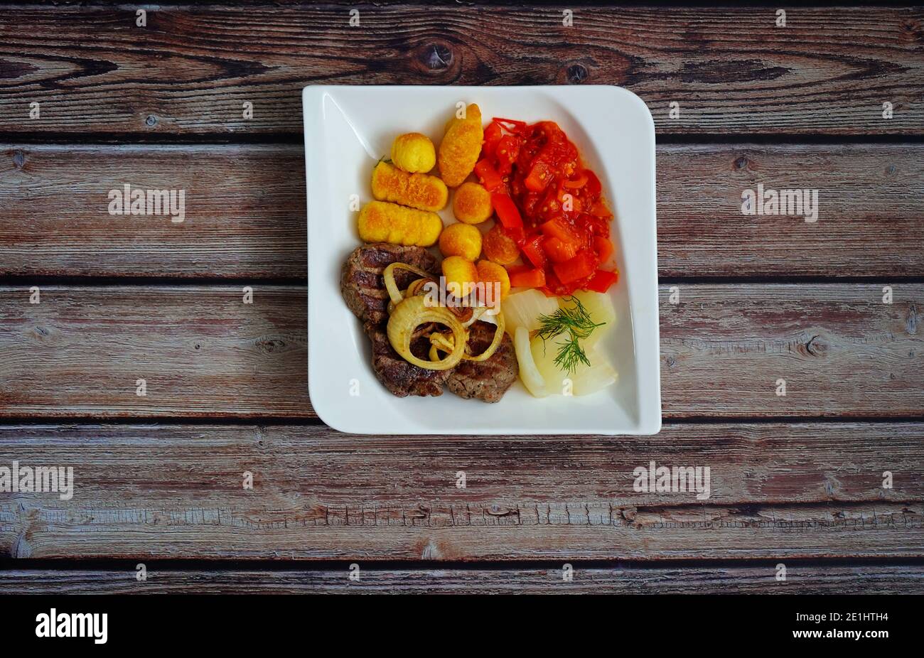 Steak de bœuf avec rondelles d'oignon, croquettes et légumes (fenouil et paprika) sur une assiette carrée blanche avec fond en bois foncé. Cuisine allemande maison. Banque D'Images