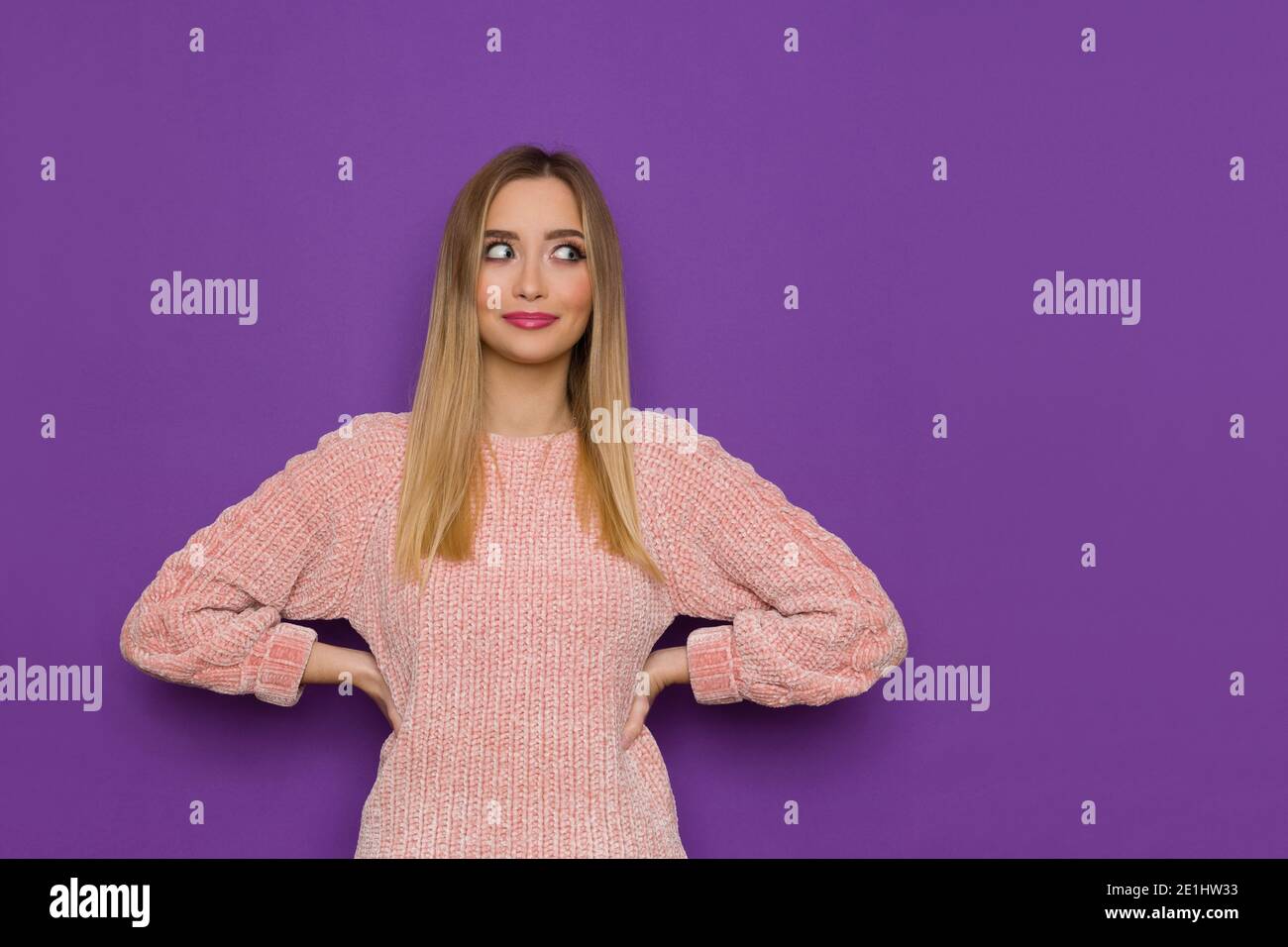 Jolie jeune femme en chandail rose pose avec les mains sur la hanche et regarde sur le côté. Taille haute, prise en studio sur fond violet. Banque D'Images