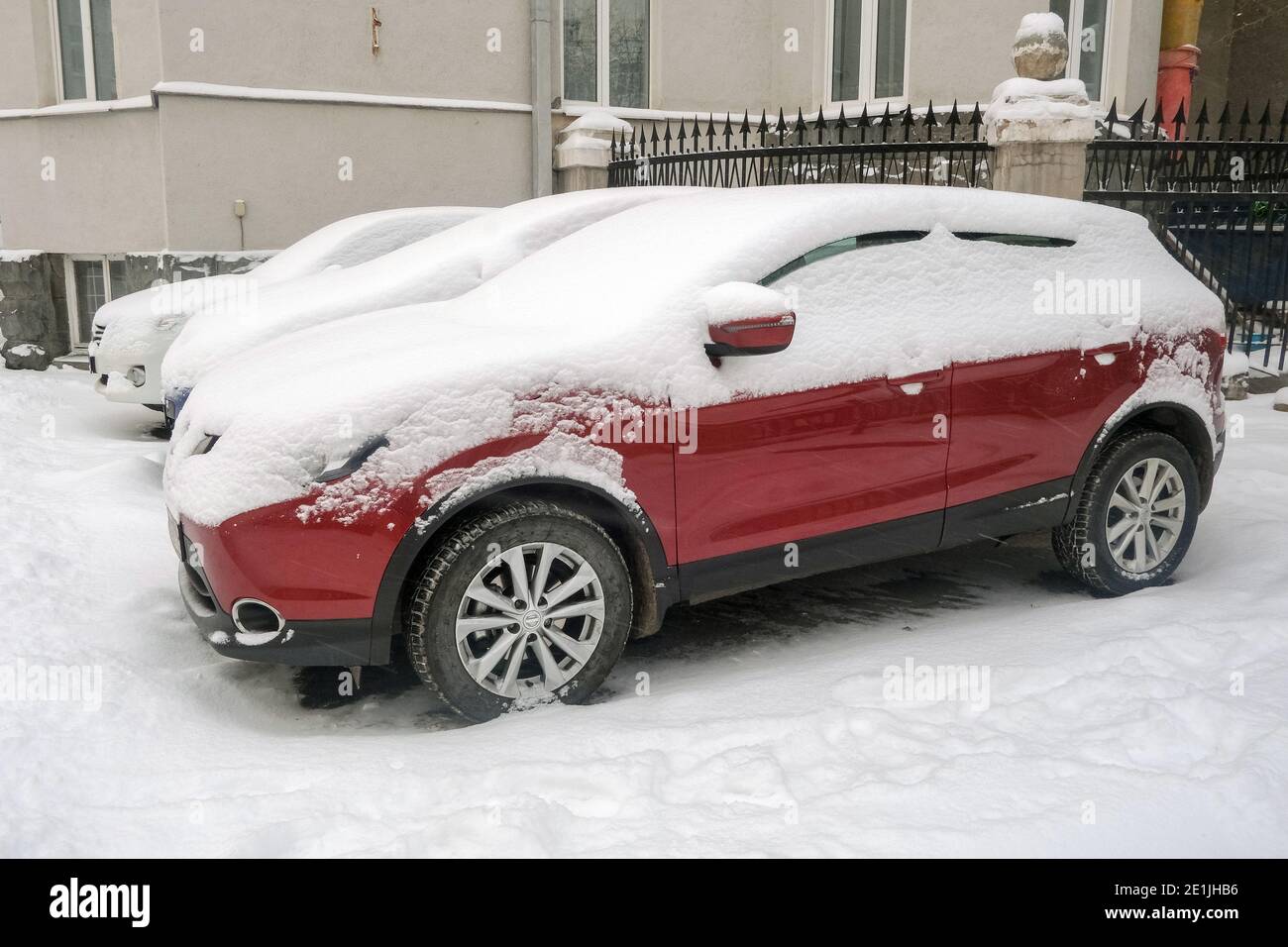 Voiture rouge moderne recouverte de neige blanche garée près de la maison dans la rue en hiver Banque D'Images