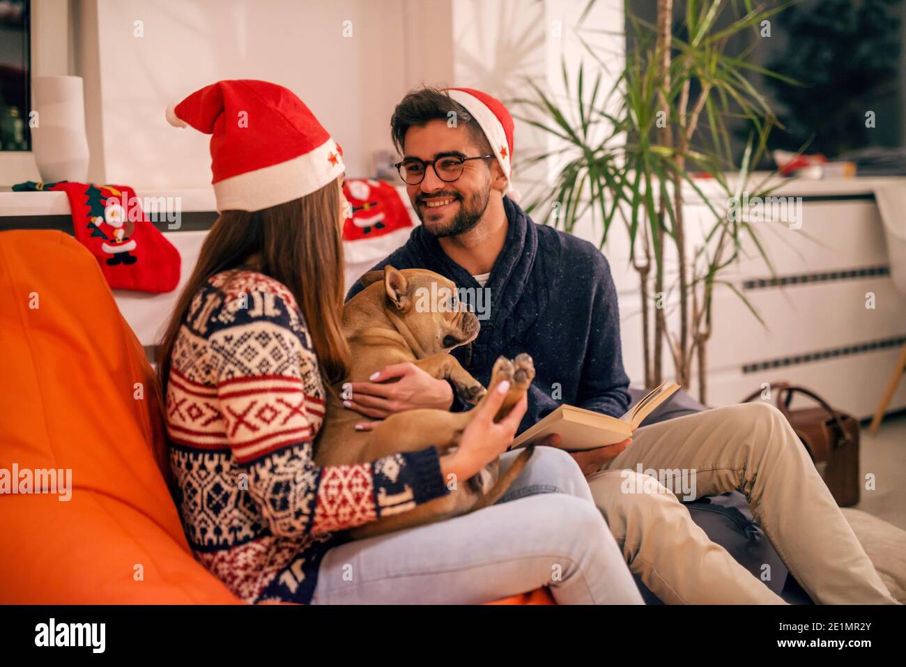 Couple passant la veille de Noël à la maison. Fille tenant chien et homme lisant un livre. Chapeau de père noël sur les têtes. Concept des fêtes de Noël. Banque D'Images