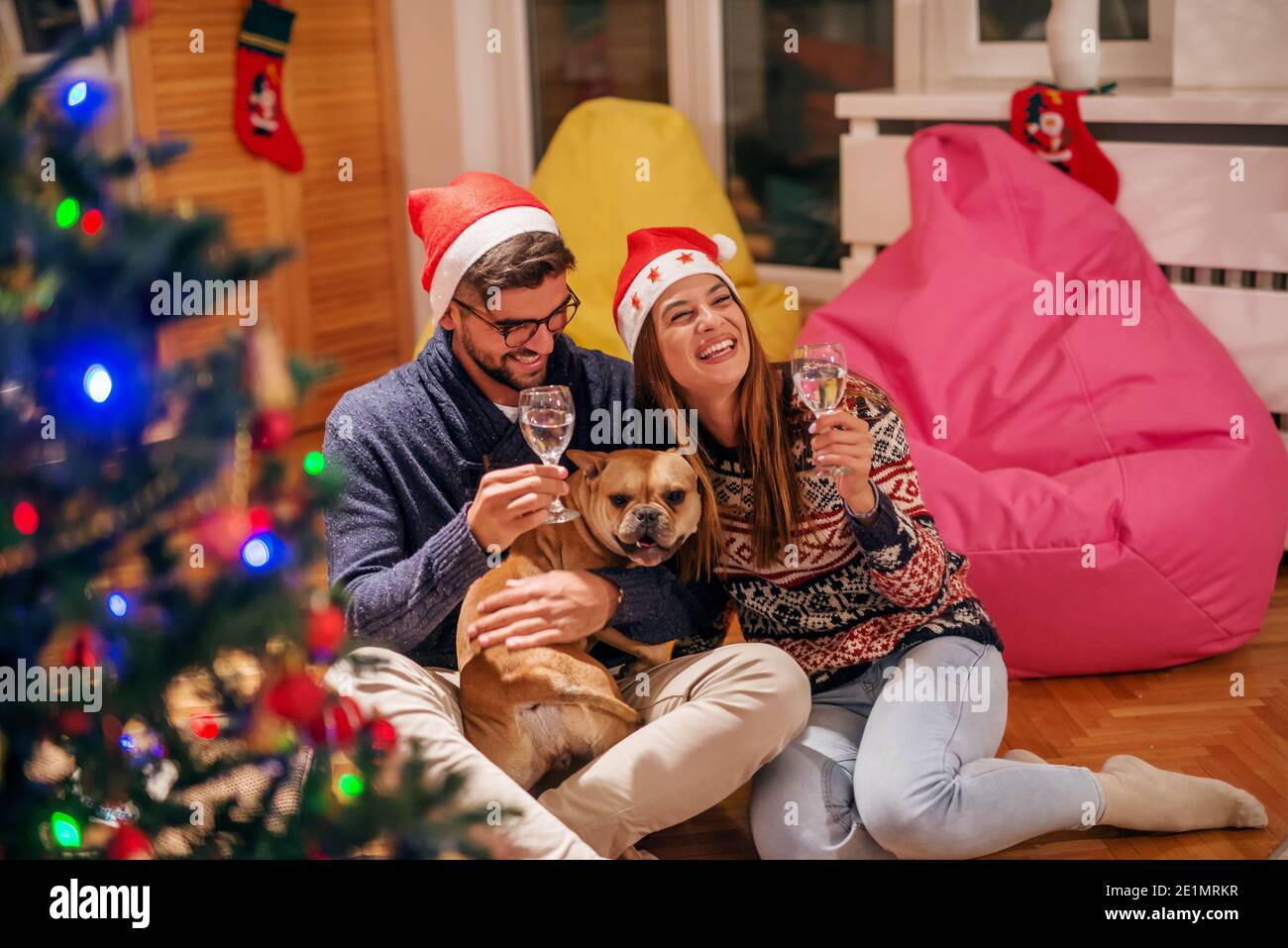 Couple assis sur le sol et buvant du champagne. Homme tenant le chien. Chapeau de père noël sur les têtes. Concept des fêtes de Noël. Banque D'Images
