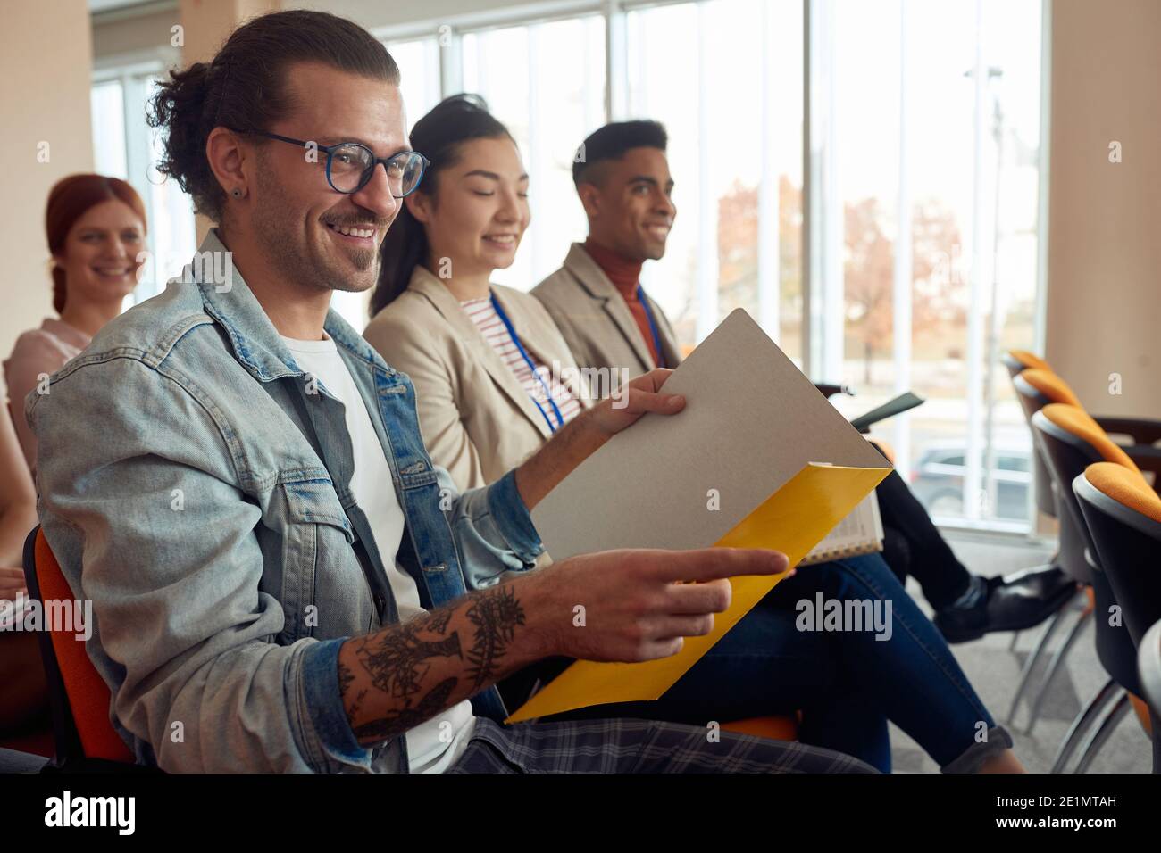 jeune homme caucasien avec tatouage portant des lunettes souriant, tenant ouvert dossier au séminaire Banque D'Images