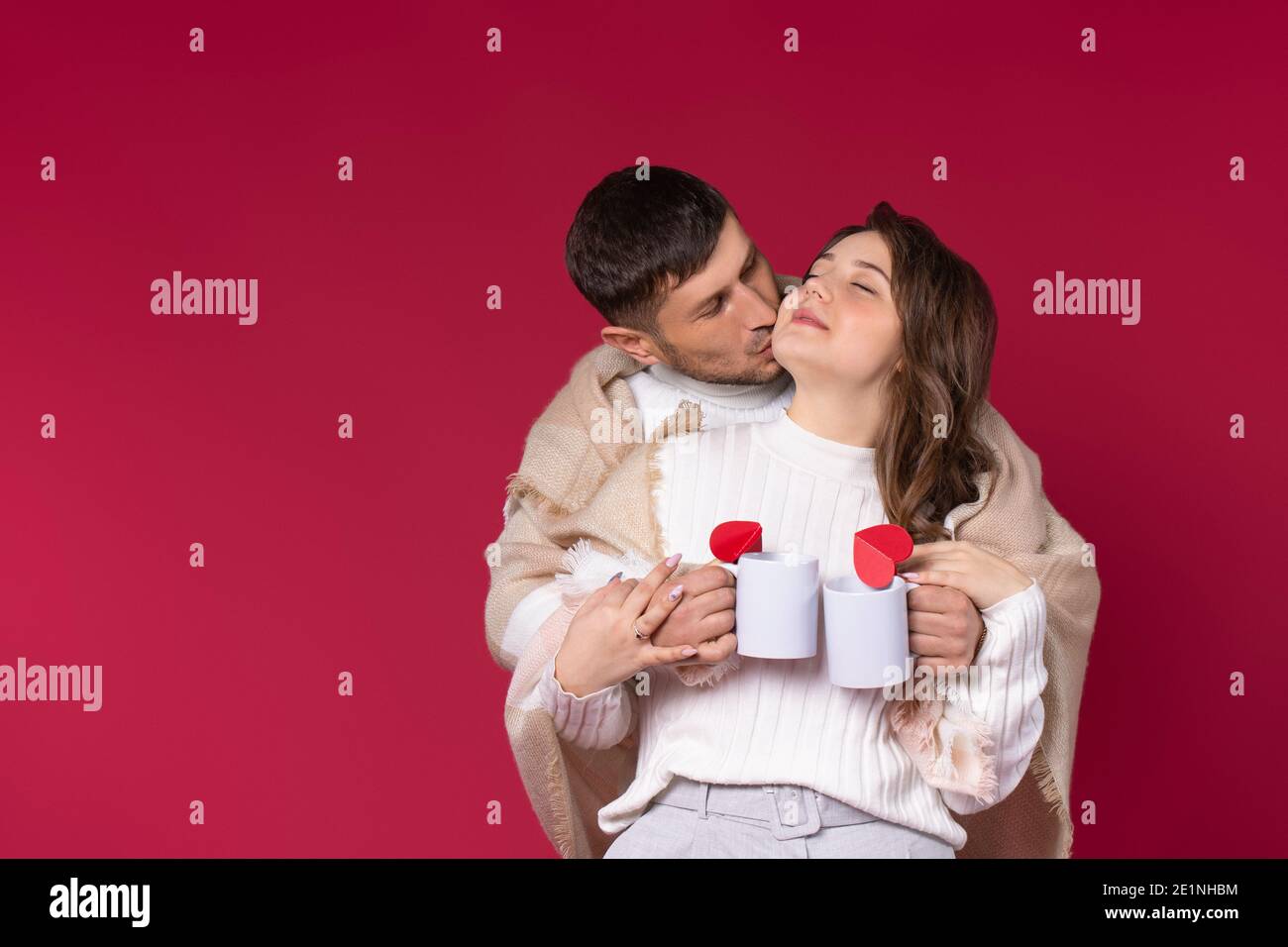 Un couple aimant enveloppé dans une couverture chaude tient les tasses à thé chaud ont un moment agréable. Arrière-plan rouge. Saint-Valentin. Banque D'Images