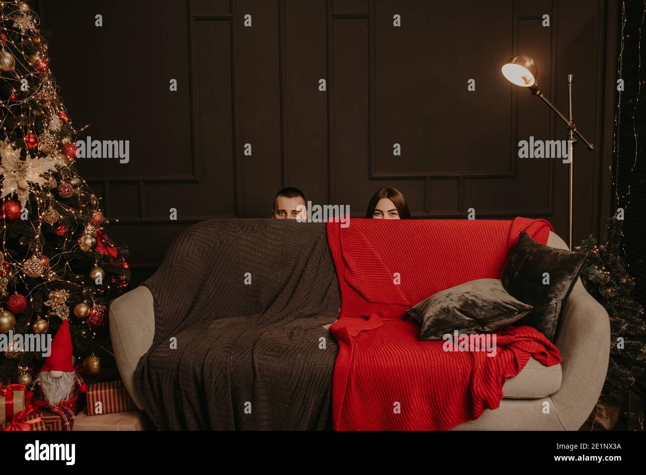 Un homme et une femme se sont cachés derrière le canapé. Les têtes d'un homme et d'une femme se tiennent derrière le canapé. Maison décorée pour le nouvel an. Le matin de Noël. Appartement dans Banque D'Images