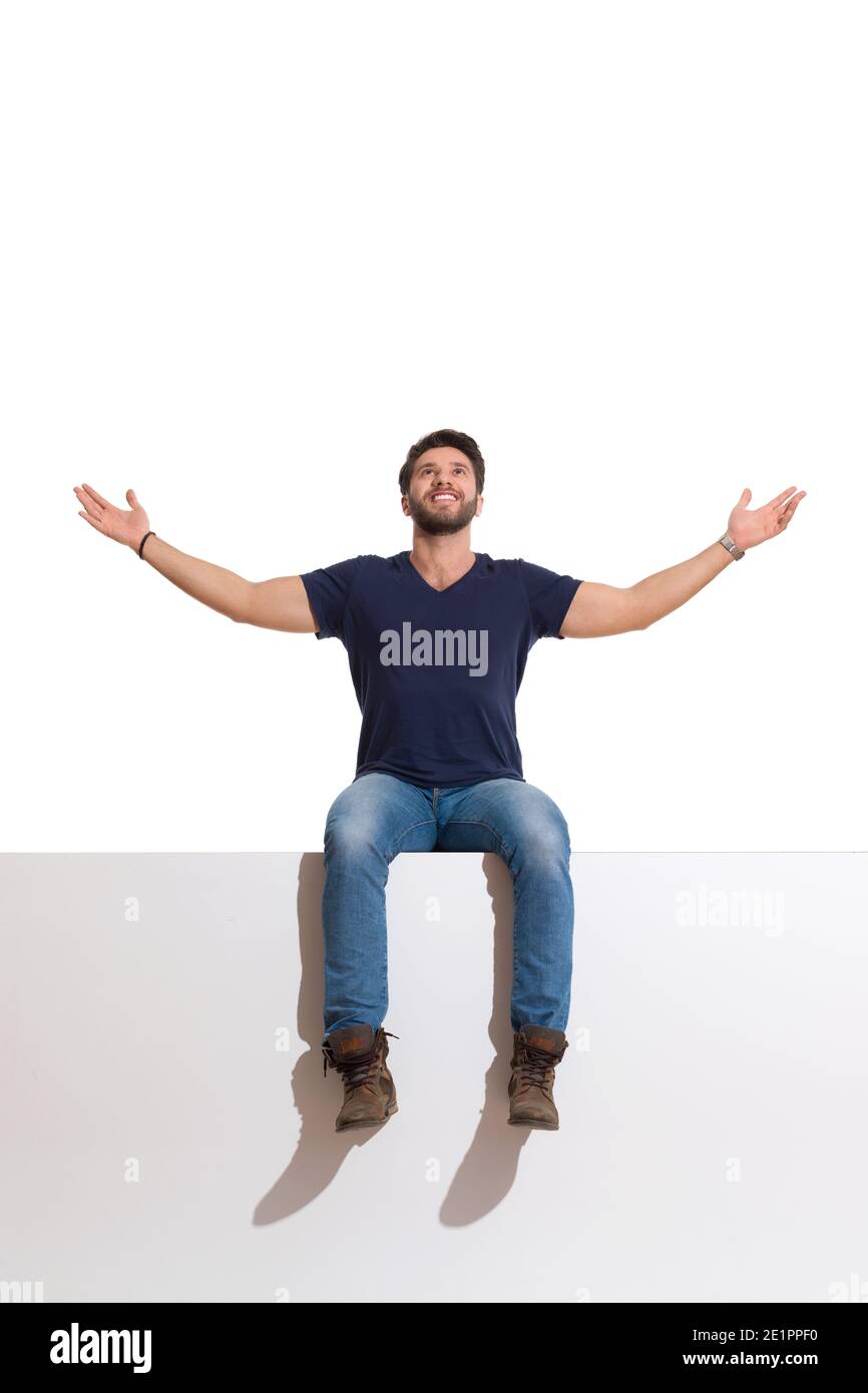 Le jeune homme heureux en bottes, jeans et chemise bleue est assis sur un haut avec les bras étirés, regardant vers le haut et souriant. Prise de vue en studio sur toute la longueur isolée sur Banque D'Images