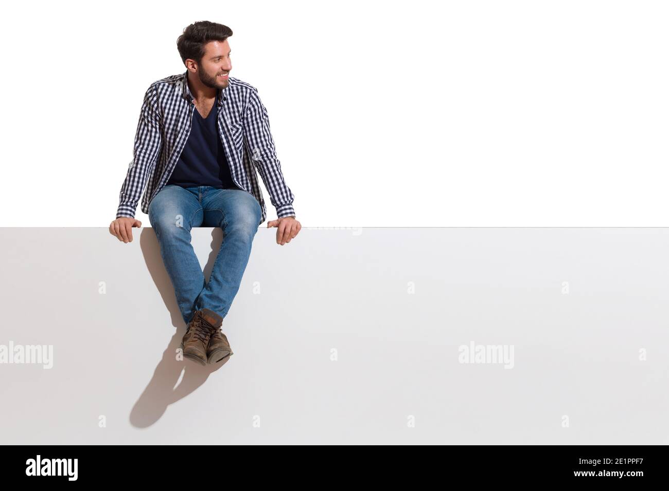 Beau jeune homme décontracté est assis sur un haut de bannière blanche avec les jambes croisées, souriant et regardant le côté. Prise de vue en studio sur toute la longueur isolée sur Banque D'Images