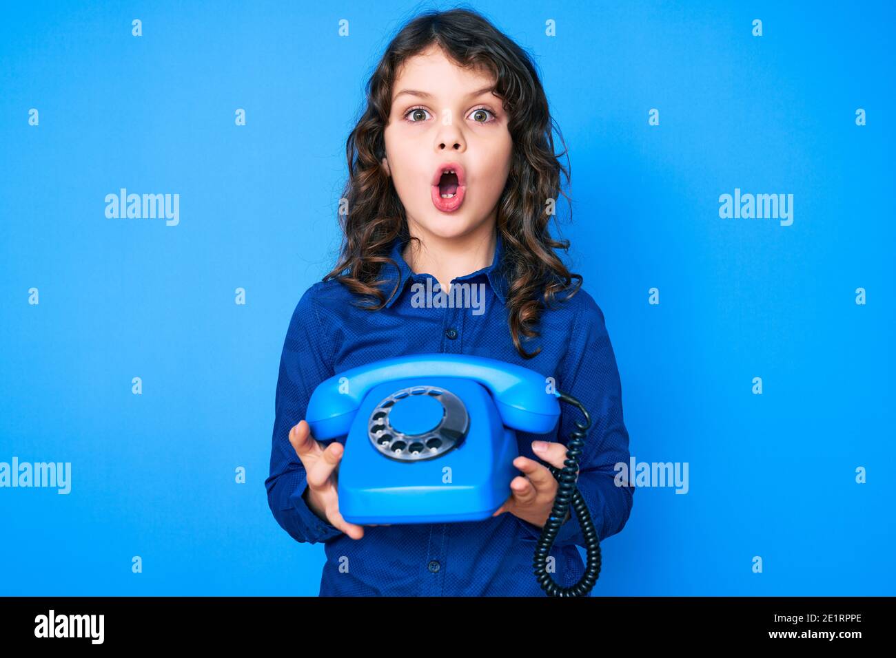 Mignon petit garçon hispanique avec de longs cheveux tenant téléphone vintage peur et choqué avec la surprise et l'expression stupéfaite, la peur et le visage excité. Banque D'Images