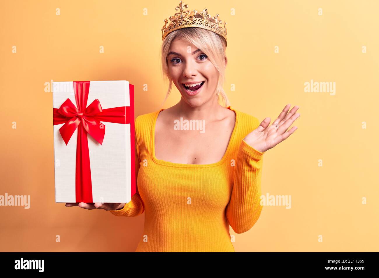Jeune belle femme blonde portant la couronne de princesse dorée tenant anniversaire cadeau célébrant la réussite avec un sourire heureux et une expression du gagnant avec r Banque D'Images Jeune belle femme blonde portant la couronne de princesse dorée tenant anniversaire cadeau célébrant la réussite avec un sourire heureux et une expression du gagnant avec r Banque D'Images