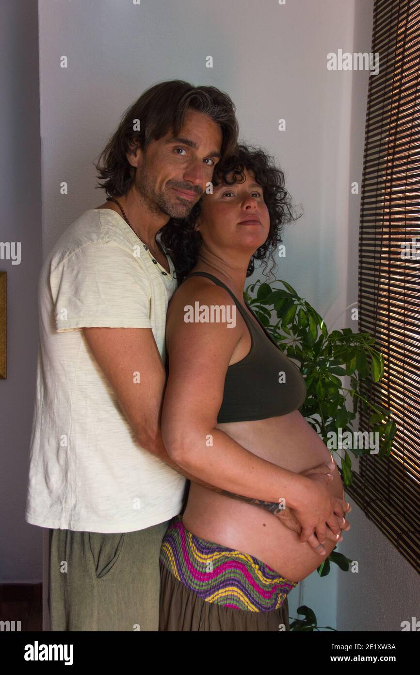 Les futurs parents debout dans la chambre à coucher se embrassant avec affection. Homme à cheveux longs tient le ventre de la femme enceinte de derrière l'intérieur. Tendresse, amour de couple Banque D'Images