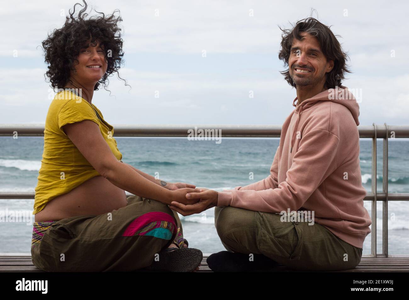 Couple souriant tenant les mains assis sur la pose de lotus sur le banc près de la plage à Gran Canaria, Espagne. Femme enceinte et futur père enyoing temps libre Banque D'Images