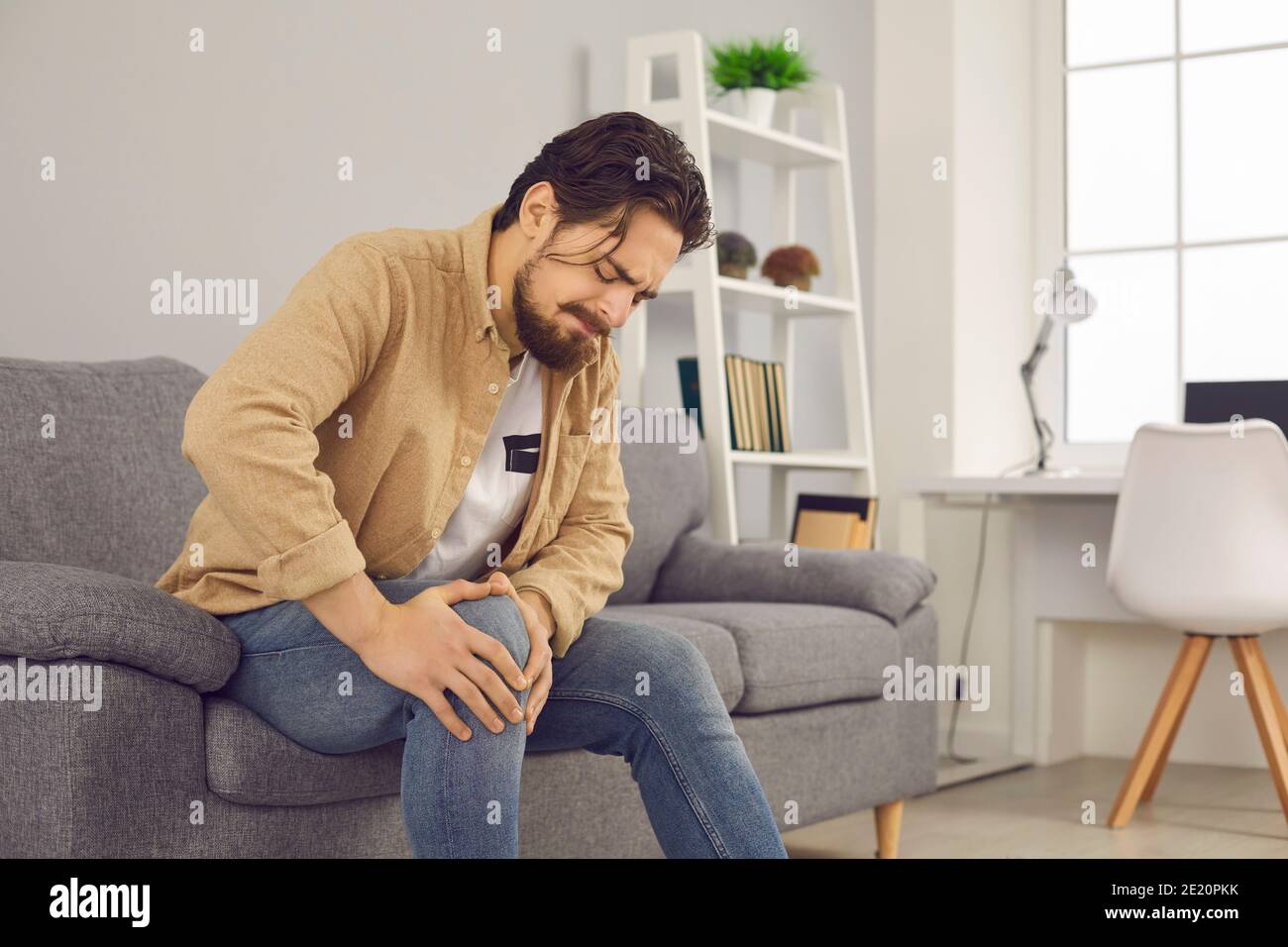 Homme assis sur un canapé à la maison et touchant le genou blessé avec la grimace de la douleur et de la souffrance Banque D'Images