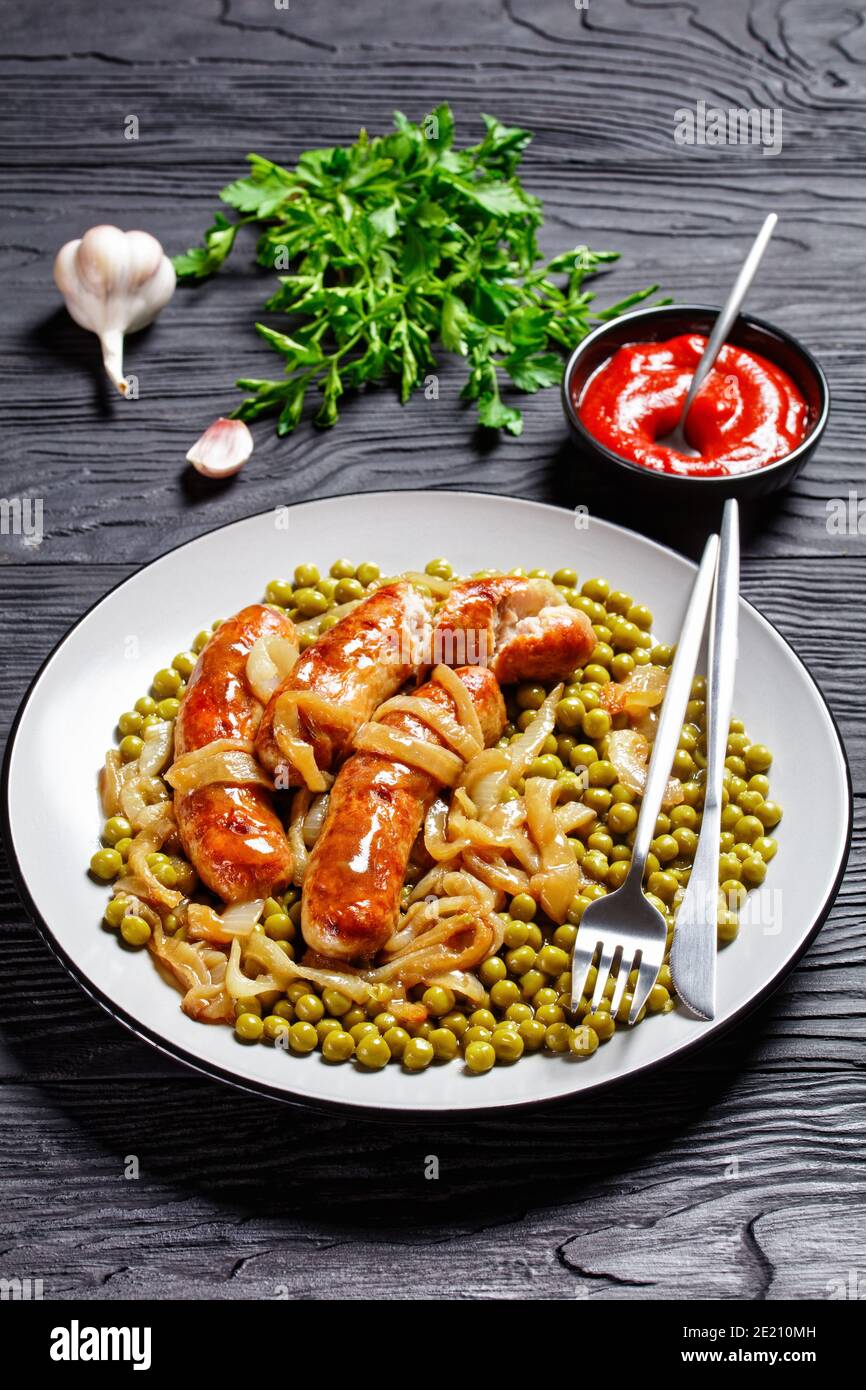 Assiette de petit-déjeuner avec saucisse de porc, petits pois verts, sauce à l'oignon et ketchup servis avec des couverts sur fond de bois sombre, vue de dessus, gros plan Banque D'Images