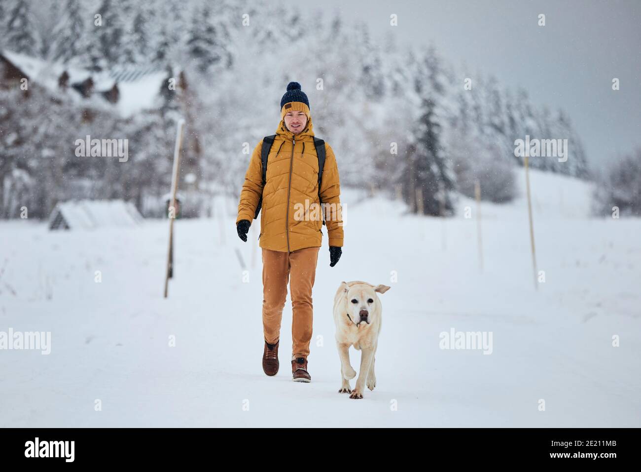 Vue avant du jeune homme avec chien en hiver. Le propriétaire d'un animal de compagnie avec son labrador retriever marchant contre la belle nature pendant le neiging. Jizera montagnes, C Banque D'Images