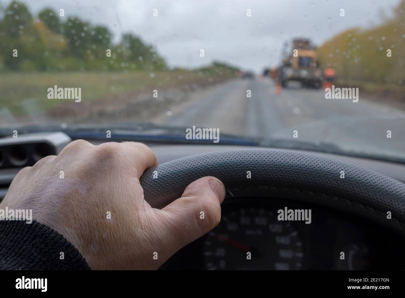 la main du conducteur sur le volant d'une voiture passant devant une section de route en réparation avec un tracteur, équipement de construction par temps pluvieux et nuageux Banque D'Images