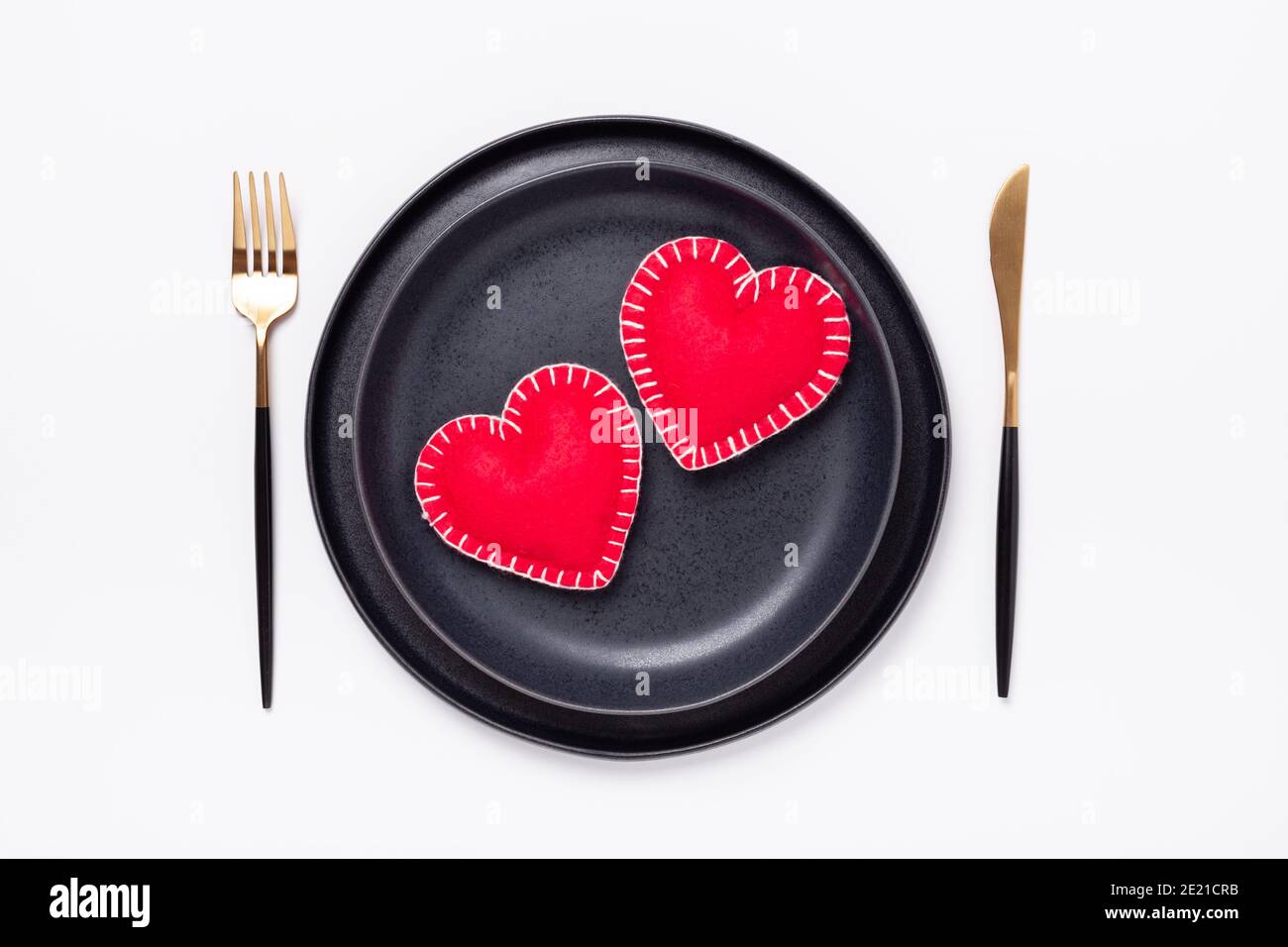 Assiette en céramique noire, fourchette et couteau, coeurs en textile rouge sur fond blanc. Table de Saint-Valentin. Vue de dessus - image Banque D'Images