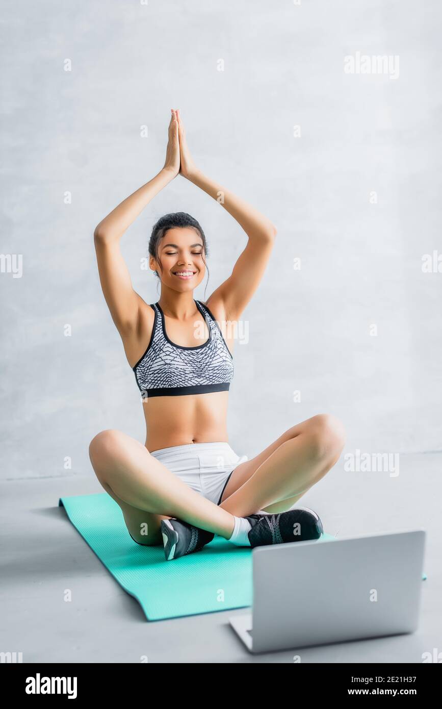 bonne femme afro-américaine assise à lotus poser avec la prière mains sur le tapis de fitness près de l'ordinateur portable Banque D'Images