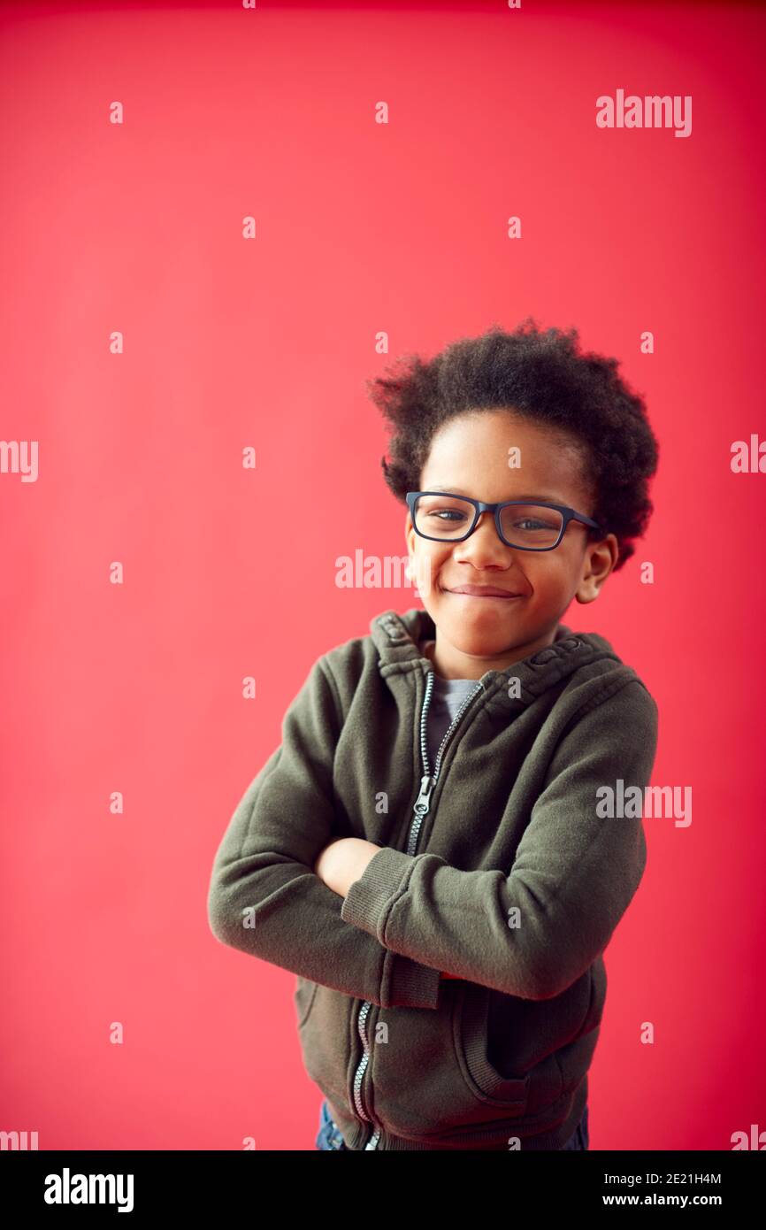 Portrait d'un garçon portant des lunettes avec des bras pliés contre le rouge Studio sourit l'arrière-plan à la caméra Banque D'Images