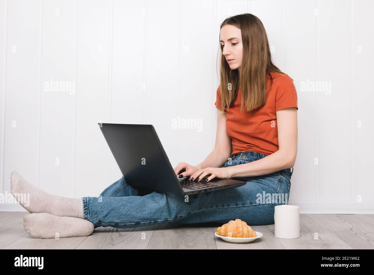 Fille souriante travaillant sur ordinateur portable tout en étant assise sur un sol avec une tasse de thé. Banque D'Images