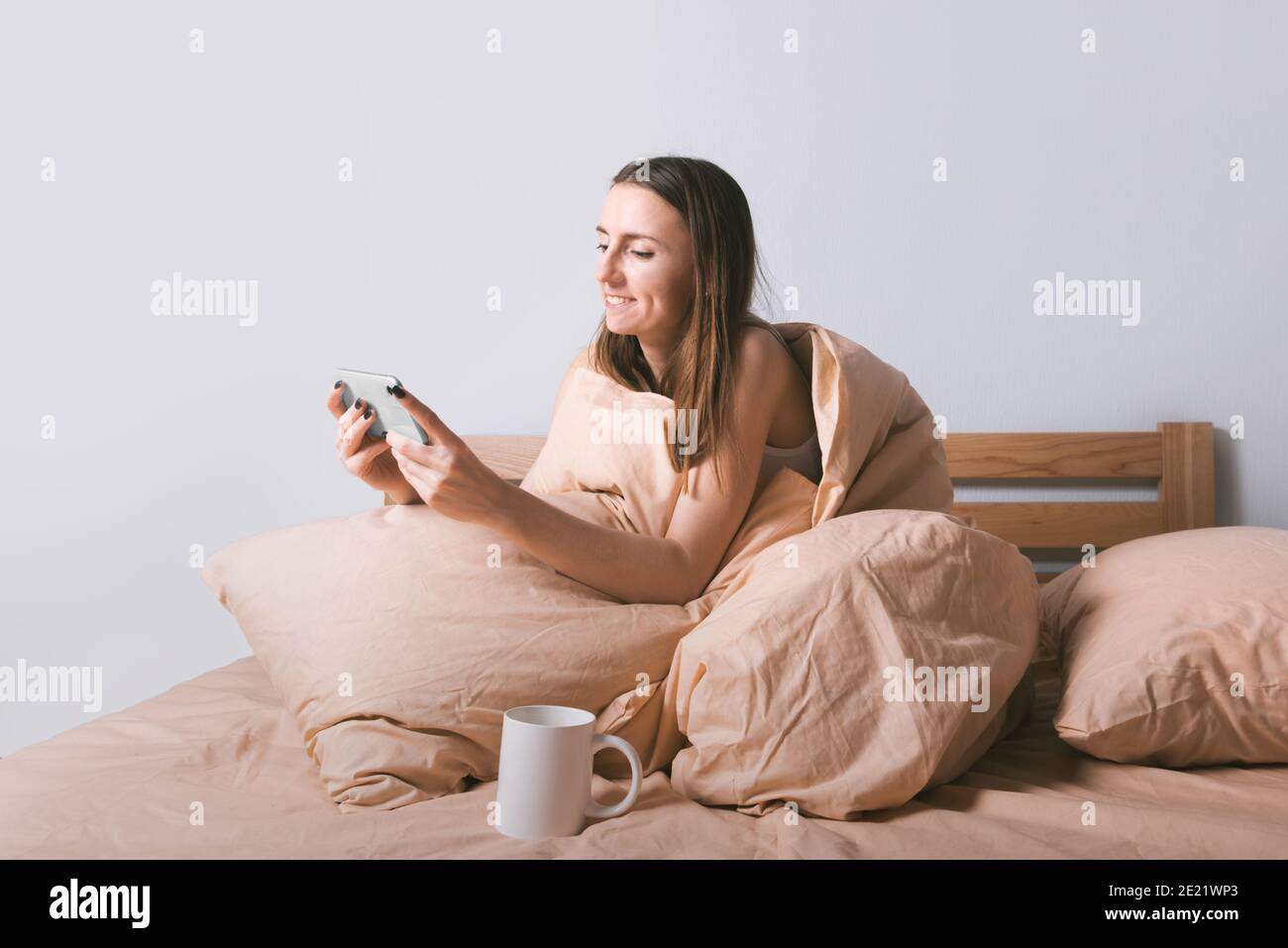 Jeune femme souriante appréciant d'utiliser un smartphone tout en étant assise dans un lit confortable le matin. Banque D'Images