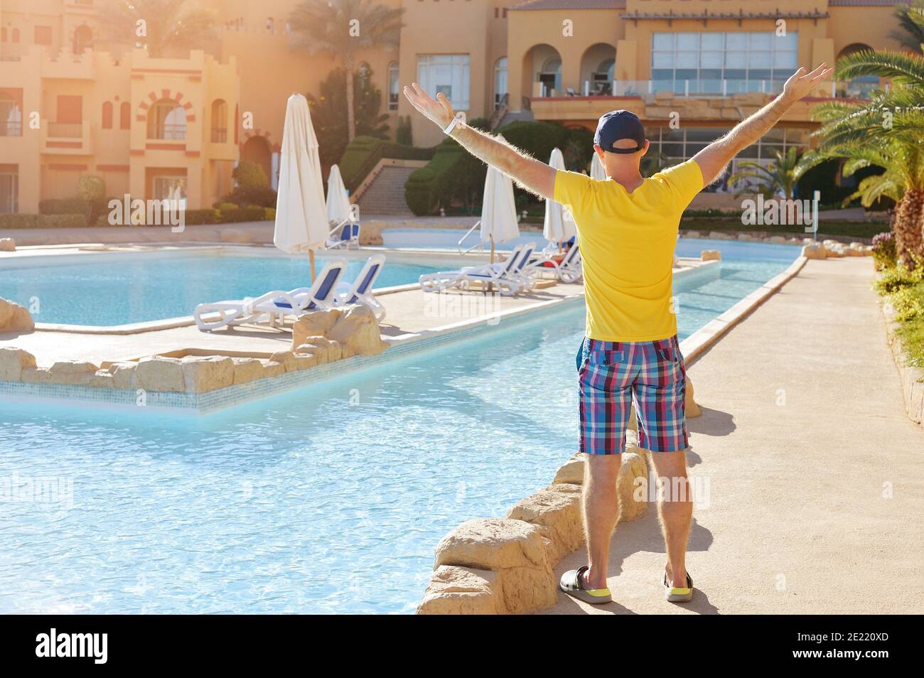 Un homme heureux lève les mains près de la piscine de l'hôtel avec un chapeau et un t-shirt jaune. Voyage, vacances d'été et concept de vacances Banque D'Images