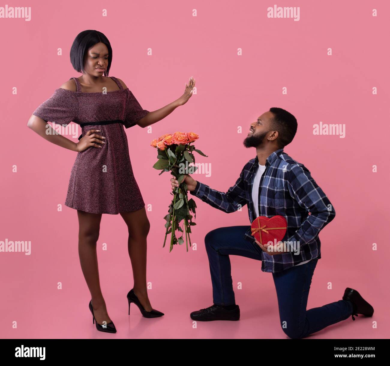 Homme noir debout sur un genou avec des fleurs et un cadeau pour la Saint-Valentin, jeune femme le rejetant sur fond rose Banque D'Images
