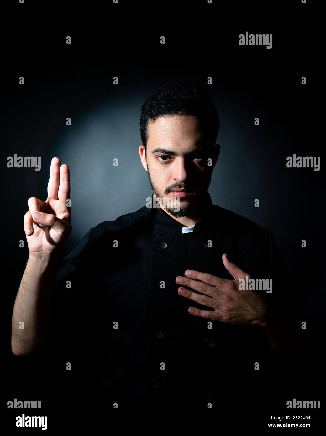 un jeune homme habillé de noir avec les vêtements du chef ressemble à un prêtre qui fait des colles. Concept spirituel. Concept de cuisine. Banque D'Images