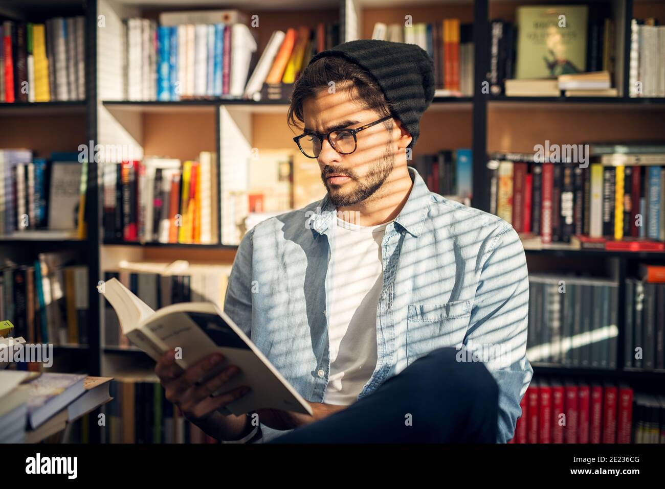 Mignon petit garçon de taille basse assis sur un plancher de bibliothèque et regardant à travers quelques livres avec le visage curieux. Banque D'Images