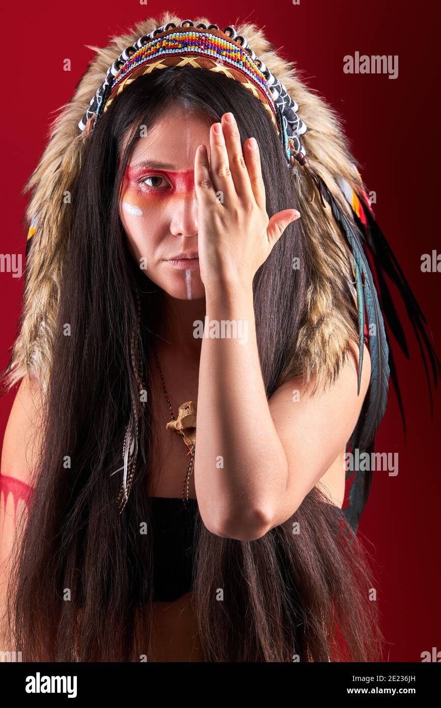 Modèle en port indien et maquillage coloré posant à la caméra, avec des plumes sur la tête. Tenue des peuples autochtones des Amériques, femme ethnique fermant h. Banque D'Images