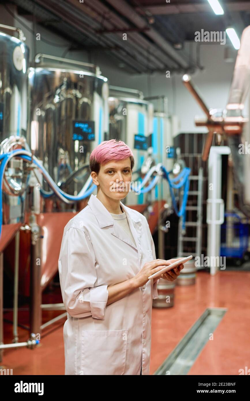Jeune femme experte en brasserie en witecavoine vous regardant pendant saisie de données dans la tablette par rapport aux nouveaux équipements pour la production de bière en usine Banque D'Images