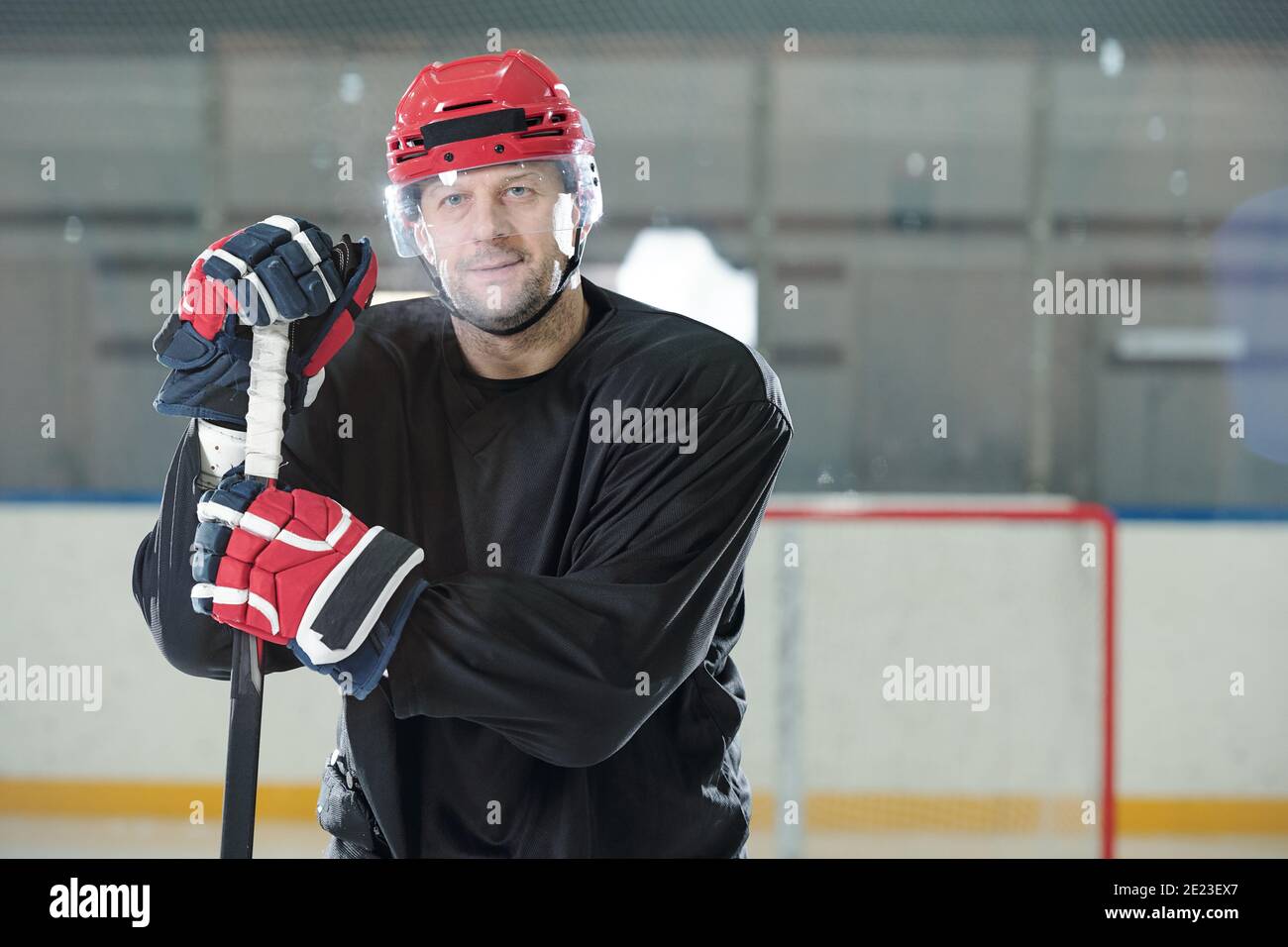 Joueur de hockey masculin mature en uniforme de sport, casque de protection et gants tenant le bâton tout en se tenant debout sur la patinoire devant la caméra et en vous regardant Banque D'Images