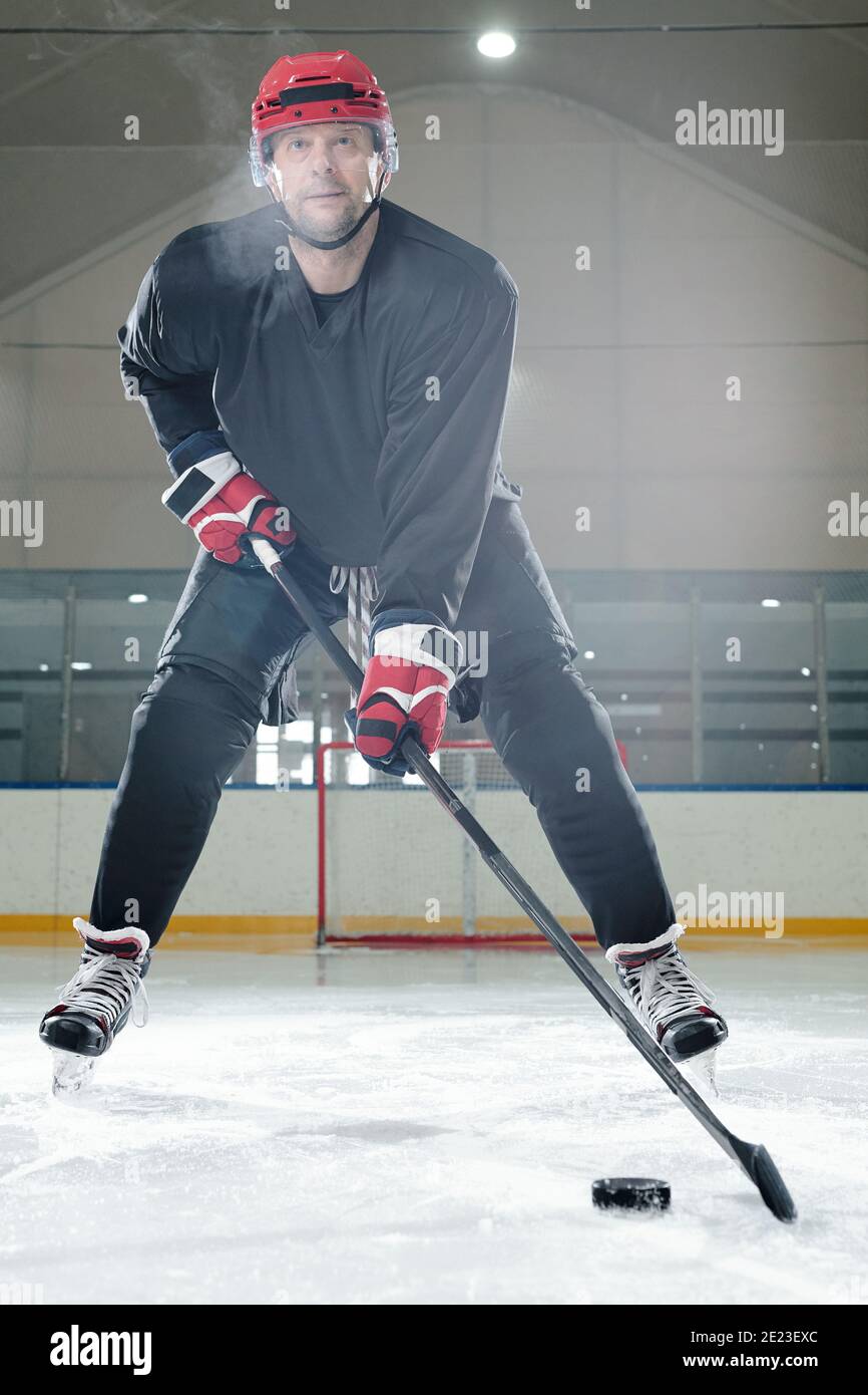 Joueur de hockey masculin mature en uniforme de sport, casque de protection et gants se déplaçant sur la patinoire devant la caméra et allant tirer sur le palet Banque D'Images