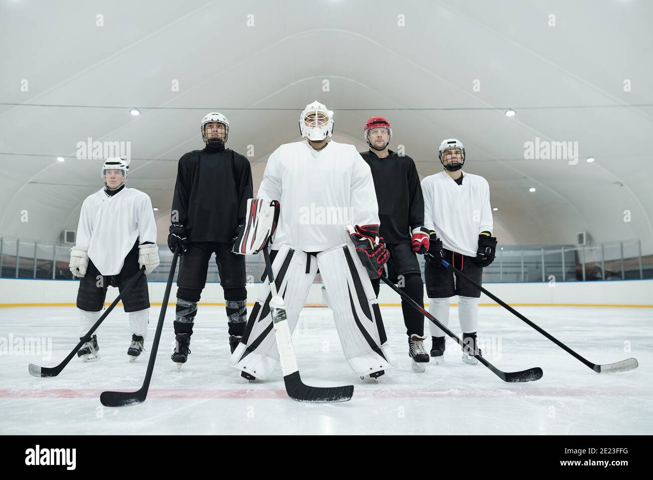 Plusieurs joueurs de hockey et leur entraîneur en uniforme sportif, gants, patins et casques debout sur une patinoire et en tenant des bâtons devant eux Banque D'Images