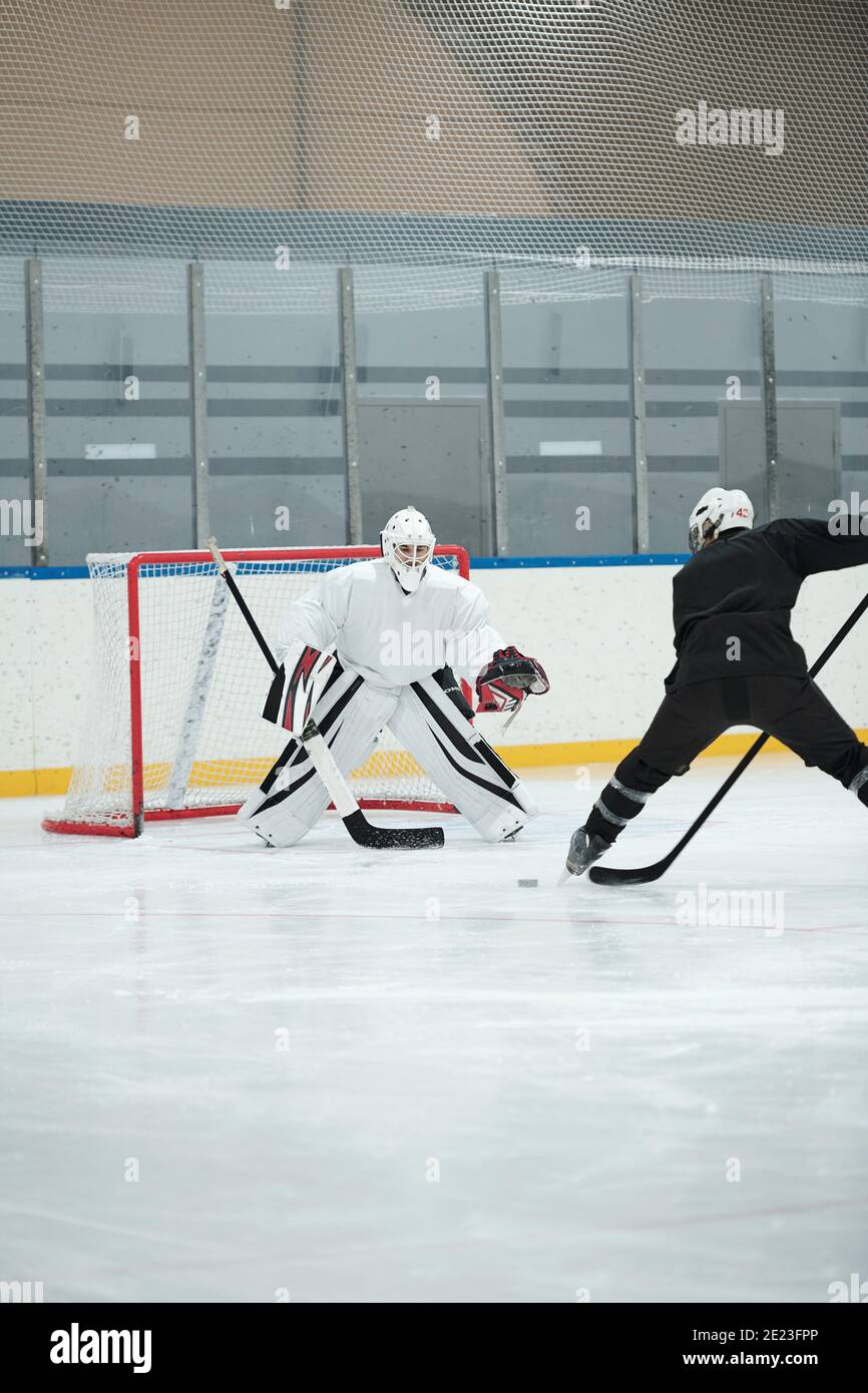 Joueur de hockey en uniforme de sport blanc, casque de protection, gants et patins debout sur la patinoire devant son rival et va tirer sur le palet pendant le jeu Banque D'Images