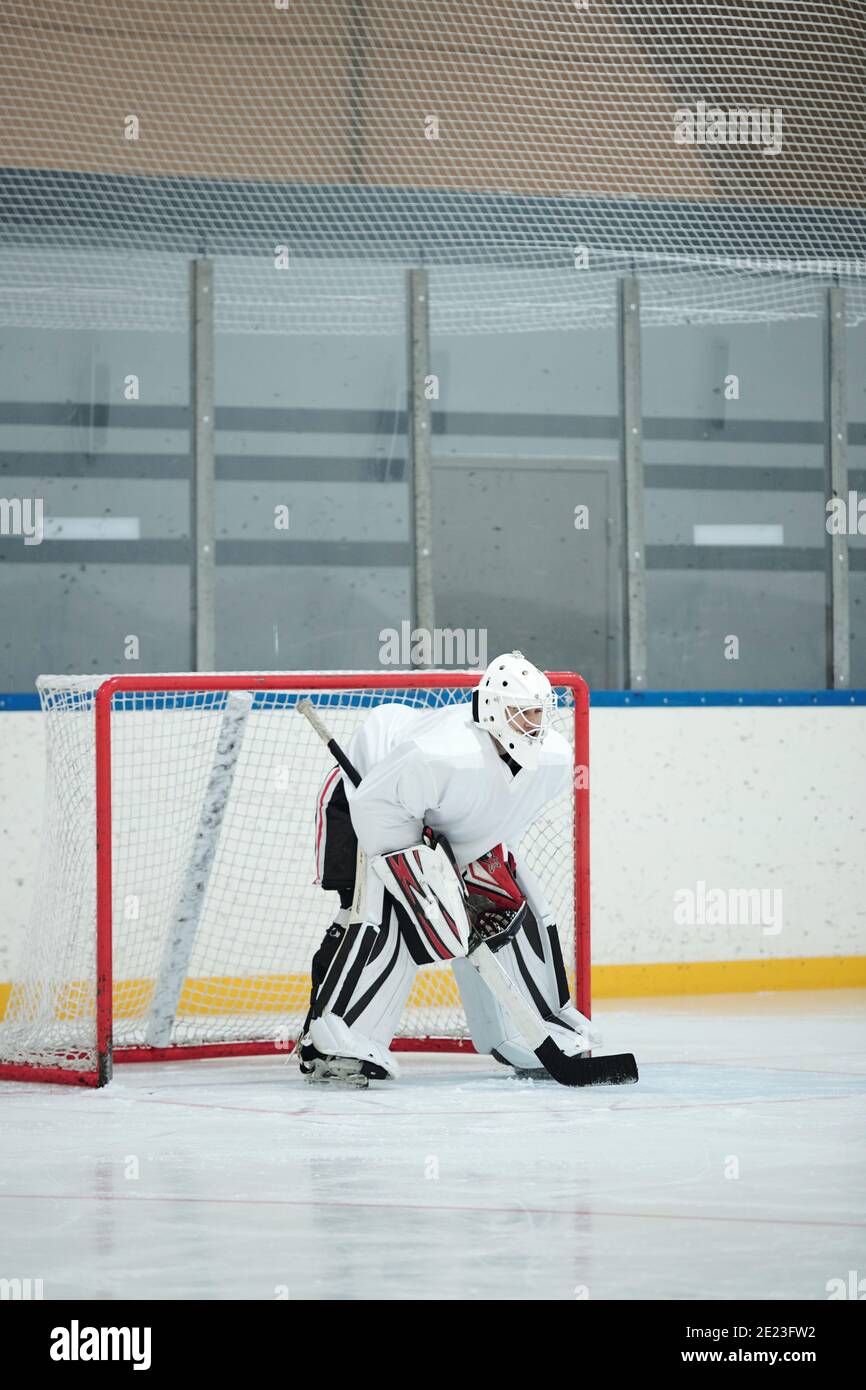 Joueur de hockey en uniforme de sport blanc, casque, gants et patins tenant le bâton tout en se pliant vers l'avant sur la patinoire devant le filet et en attendant le palet Banque D'Images
