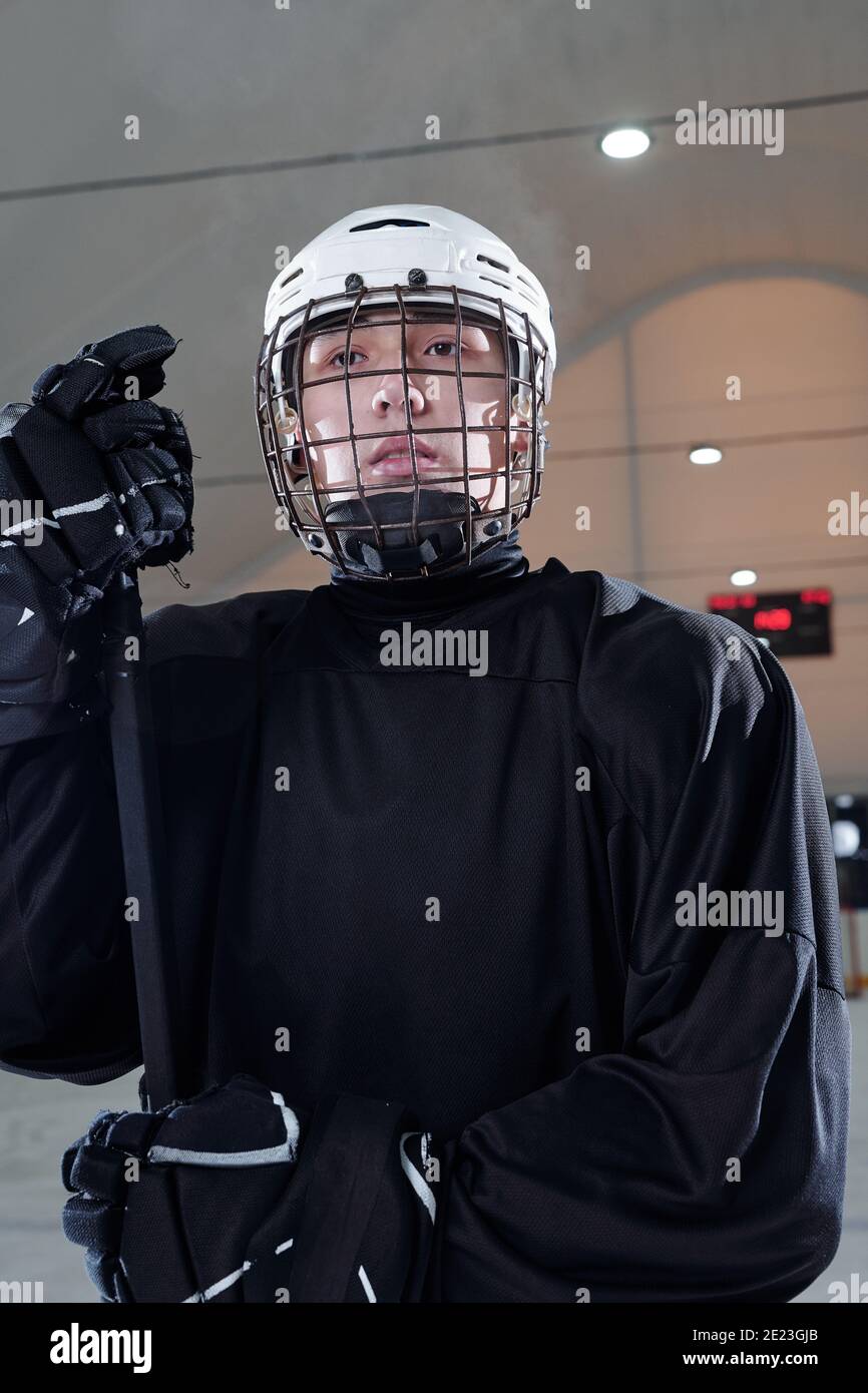 Jeune joueur de hockey contemporain en uniforme sportif noir, gants et casque de protection regardant vers l'avant en se tenant debout sur la patinoire Banque D'Images