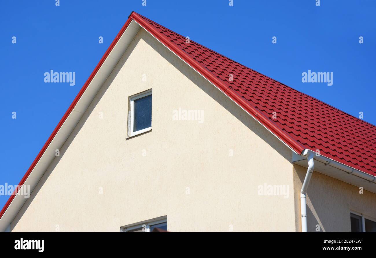 Un gros plan sur un toit en métal rouge et un soffit en plastique d'une maison avec des murs en stuc, une petite fenêtre de grenier et un système de gouttière de toit. Banque D'Images