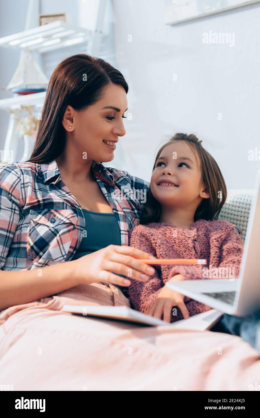 Mère souriante regardant la fille tout en pointant avec un crayon à ordinateur portable flou au premier plan Banque D'Images